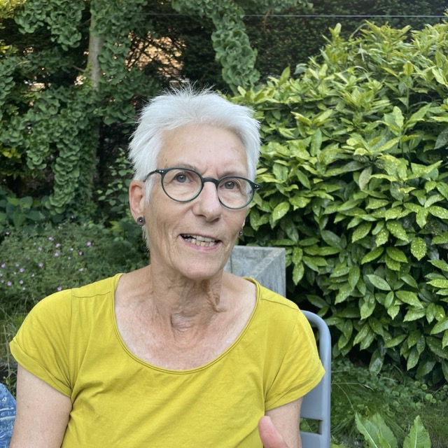 Portret van oudere vrouw met bril en geel shirt buiten.