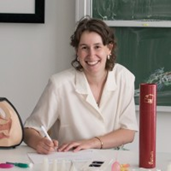 Vrouw achter bureau met boek en pen, lachend naar camera.